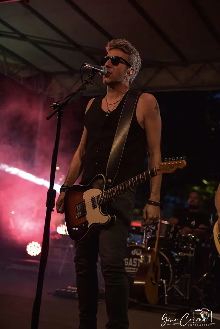 Il cantante in canottiera nera canta al microfono sul palco, con la batteria alle spalle
