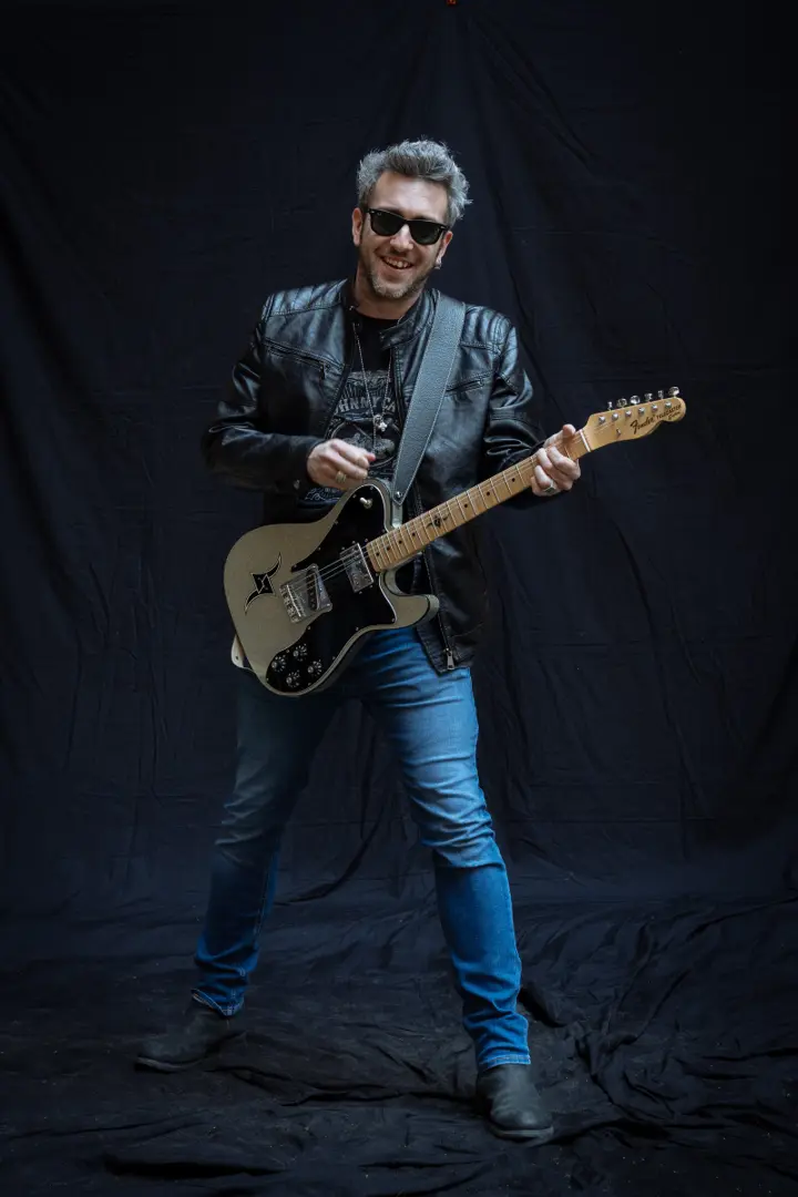 Nazareno Albanesi dei Souvenirs sorridente, in giacca di pelle e jeans, con la chitarra elettrica su sfondo nero